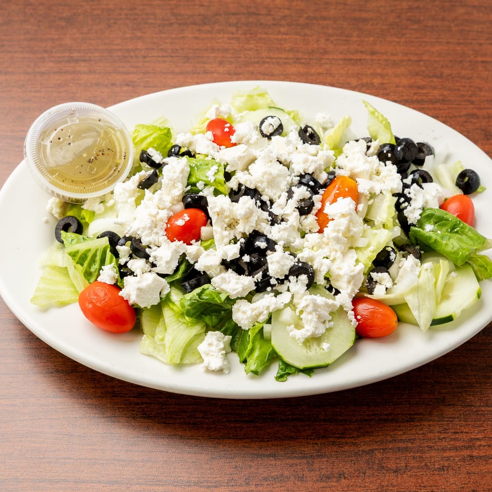 Greek salad with lettuce, tomatoes, cucumbers, black olives, crumbled feta, and dressing on a white plate.